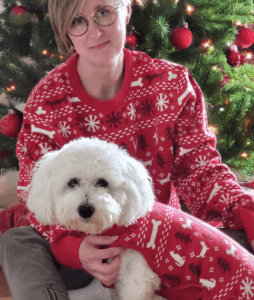 En kvinna i röd tröja håller en vit hund framför en julgran.