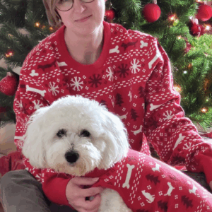 En kvinna i röd tröja håller en vit hund framför en julgran.