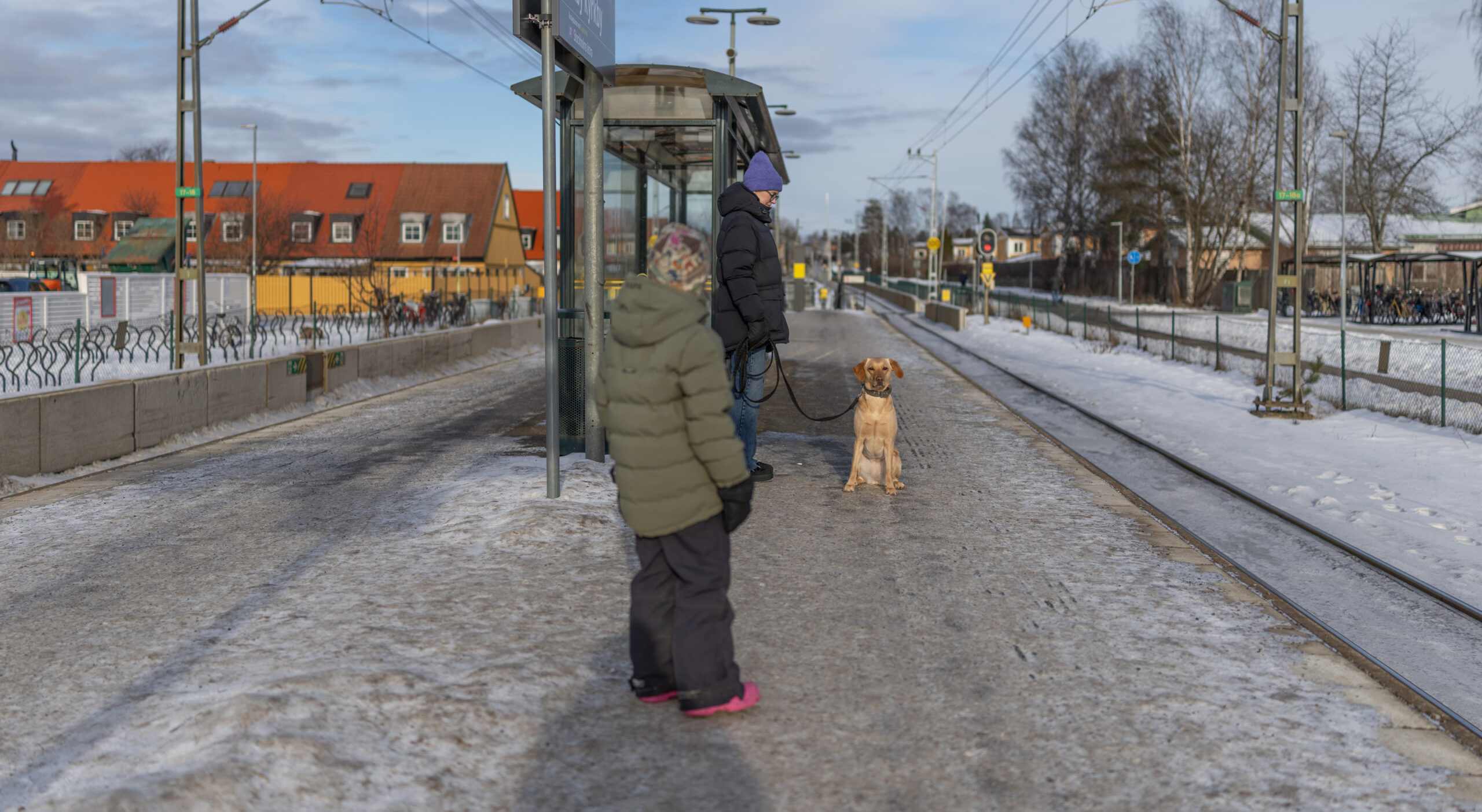 En person står bredvid sin hund på en tåg station, ett barn står i närheten och tittar på hunden.