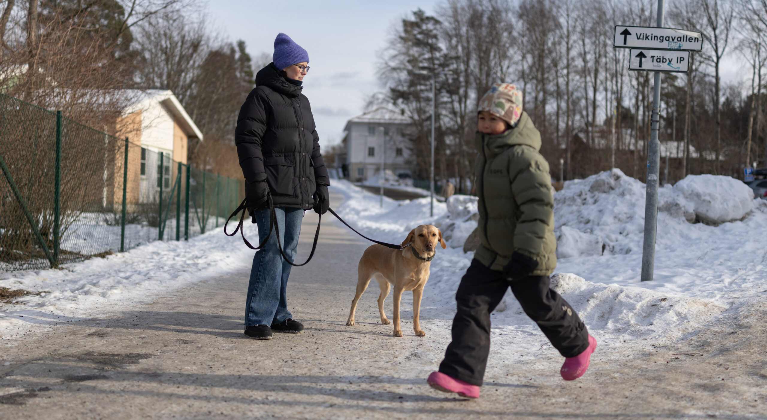 En person står på en snötäckt gata bredvid sin hund, ett barn passerar.